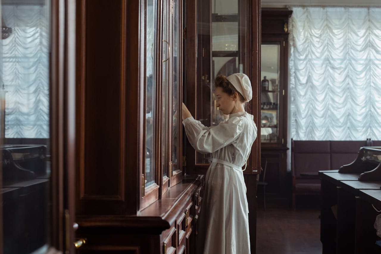 A woman in a vintage white dress exploring wooden cabinets in a timeless library interior.